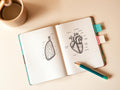 Anatomy notebook with hand-drawn heart and lung diagrams, washi-tape tabs, and a pencil on a cream desk